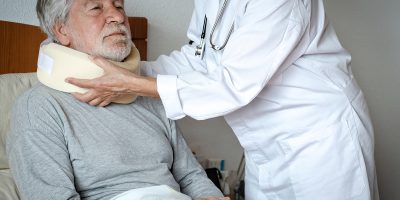 Senior bearded man in pain wearing a cervical collar with the assistance of a physiotherapist doctor during a visit home